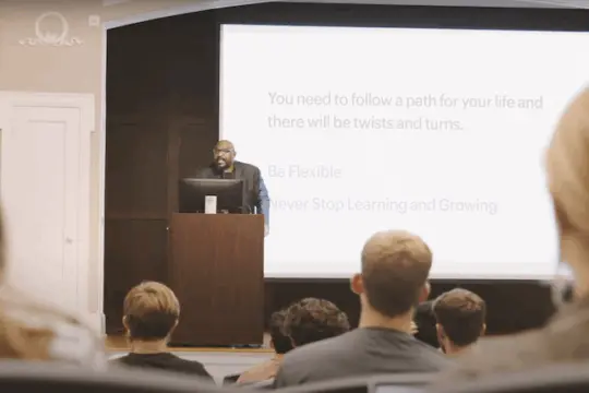 A speaker stands at a podium in front of a classroom, addressing students. Behind him is a large screen displaying a slide with the text: 